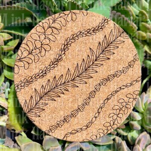 May include: A round cork coaster with a natural brown background. The coaster features four diagonal designs in black, including floral and leaf patterns. The coaster is surrounded by green succulent plants.
