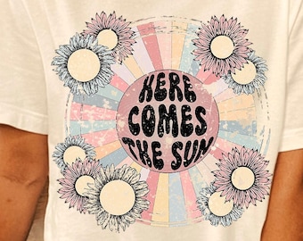 May include: White t-shirt with a graphic print of a rainbow sun with the text "Here Comes The Sun" and pink and blue flowers.