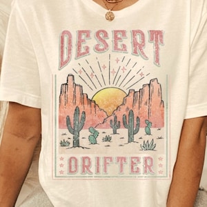 May include: White t-shirt with a graphic print of a desert scene with mountains, a sunset, and cacti. The text "DESERT DRIFTER" is printed in pink on the shirt.