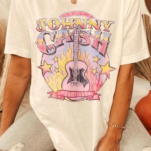 May include: White t-shirt with a graphic print of a guitar and the text "Johnny Cash" in a colorful vintage style. The graphic also includes the text "Nashville" in a banner.