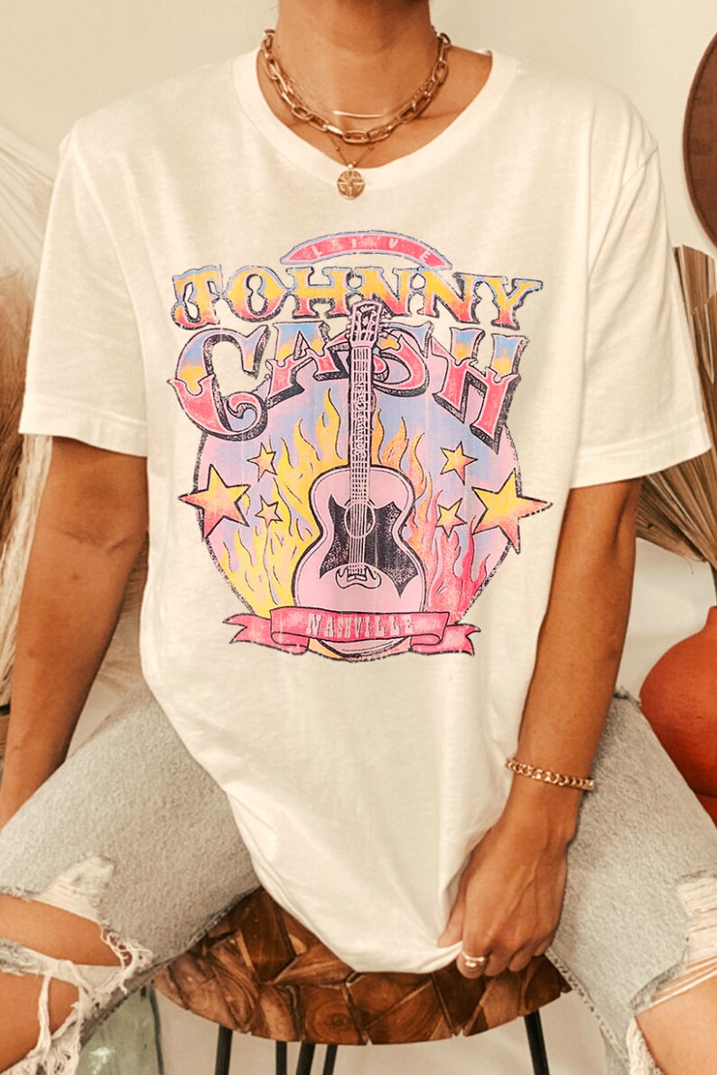 May include: White t-shirt with a graphic print of a guitar and the text "Johnny Cash" in a colorful vintage style. The graphic also includes the text "Nashville" in a banner.