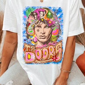 May include: White t-shirt with a colorful graphic print of Jim Morrison, the lead singer of The Doors. The graphic features a psychedelic design with flowers, swirls, and the band's name.