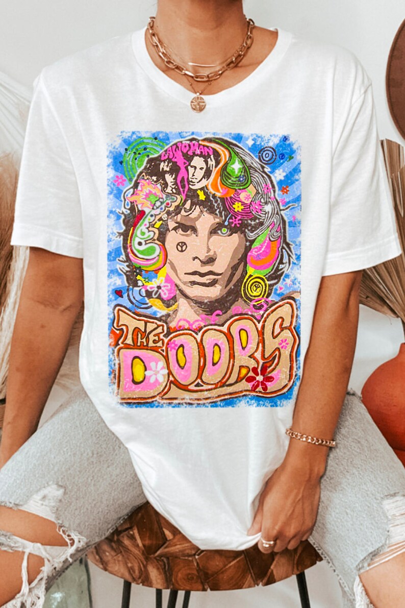 May include: White t-shirt with a colorful graphic print of Jim Morrison, the lead singer of The Doors. The graphic features a psychedelic design with flowers, swirls, and the band's name.