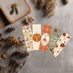 Può includere: Cinque segnalibri decorativi con disegni a tema autunnale. I segnalibri presentano zucche, tazze di caffè e motivi a quadri arancioni, verdi e marroni. Il testo include "In My Cozy Girl Era" e "Cozy Book Club."