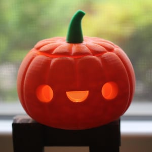 Puede incluir: Una calabaza naranja sonriente con un tallo verde, perfecta para Halloween. La calabaza tiene dos agujeros redondos para los ojos y una boca curva, con una luz cálida en el interior. Se encuentra sobre una superficie oscura.