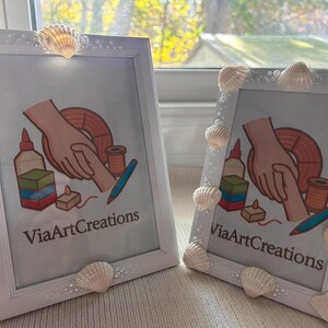 May include: Two white picture frames decorated with seashells. Each frame displays a colorful illustration with the text "ViaArtCreations". The frames are leaning against a window.