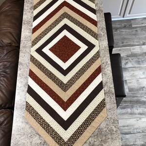 May include: A geometric table runner with a diamond pattern. The runner features alternating stripes of brown, beige, and white fabrics. The central diamond is a darker brown, and the edges are a lighter beige.