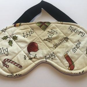 May include: Quilted eye mask with a cream background and a black strap. The mask features a repeating pattern of Christmas-themed illustrations, including holly, gifts, and a bird. The words "merry", "snow", "gift", and "bird" are also printed on the mask.