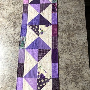 May include: A handmade, quilted table runner in shades of purple, lavender, and cream. The design features geometric shapes, including triangles and squares, with floral and polka dot patterns. The runner is approximately 36 inches long.