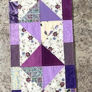 May include: Purple and white patchwork table runner with a floral pattern. The runner is made of different fabrics and is sewn together in a geometric pattern.
