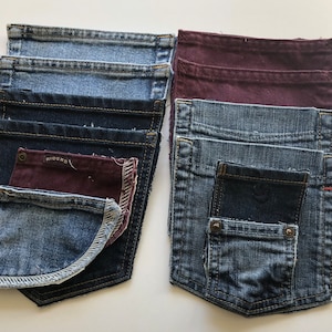 May include: A collection of denim pockets in various shades of blue and burgundy. The pockets are arranged in two stacks, showcasing different washes and styles. Some pockets have visible stitching and rivets, while others feature the "RIDERS" brand label.