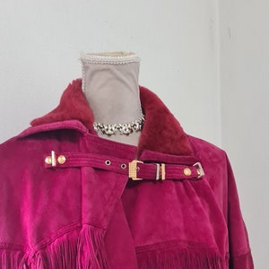 May include: A long, burgundy suede jacket with fringe detailing and gold hardware. The jacket has a fur collar and a belted waist.