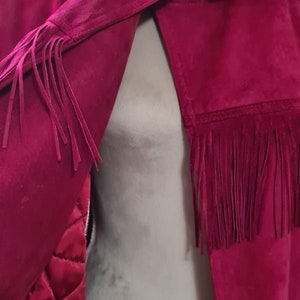 May include: A burgundy suede jacket with a fur collar and fringe detailing. The jacket has a gold buckle closure and a quilted lining.