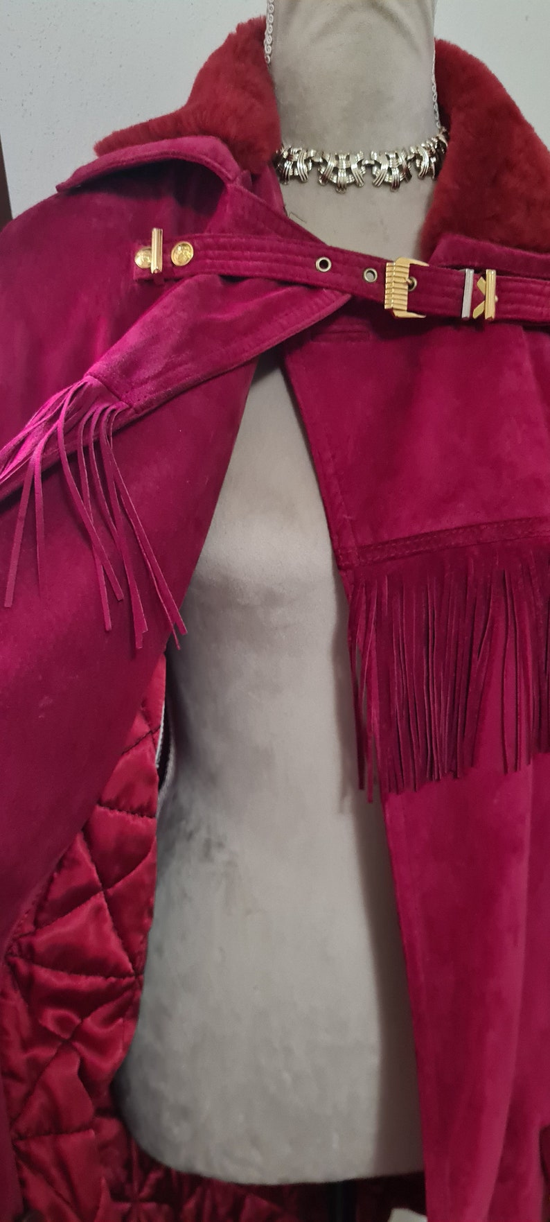 May include: A burgundy suede jacket with a fur collar and fringe detailing. The jacket has a gold buckle closure and a quilted lining.