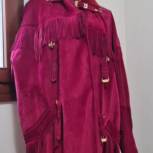 May include: A long, burgundy suede jacket with fringe detailing and gold-toned hardware. The jacket has a collar and a front closure.