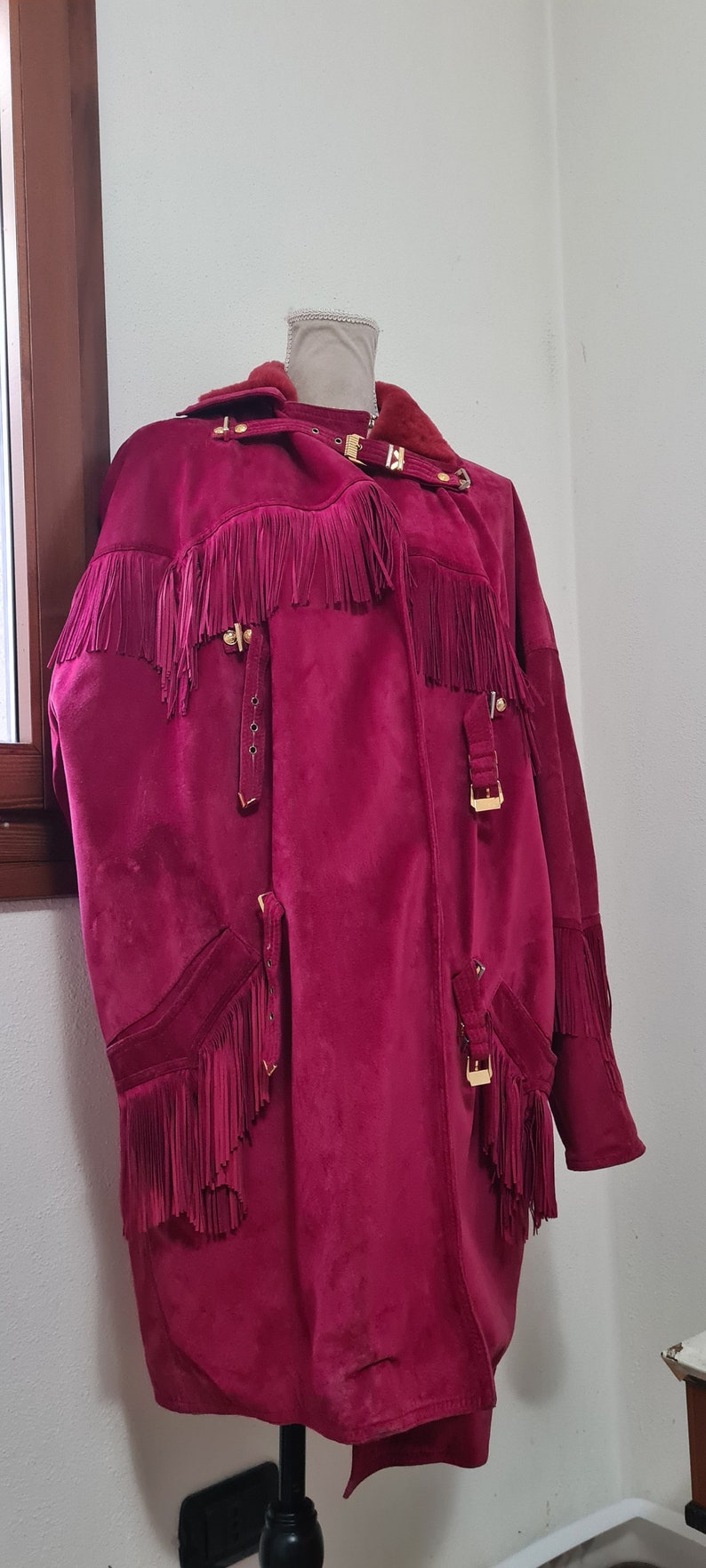 May include: A long, burgundy suede jacket with fringe detailing and gold-toned hardware. The jacket has a collar and a front closure.