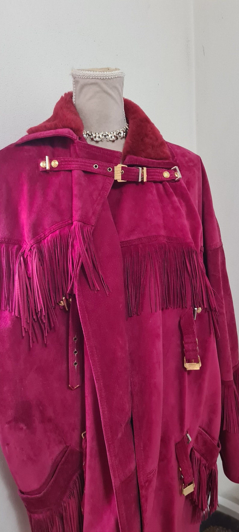 May include: A long, burgundy suede jacket with fringe detailing and gold hardware. The jacket has a fur collar and a belted waist.