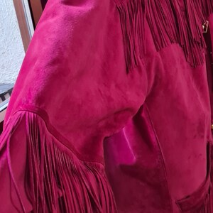 May include: A pink suede jacket with fringe detailing. The jacket has a gold buckle on the collar and a pocket on the front.