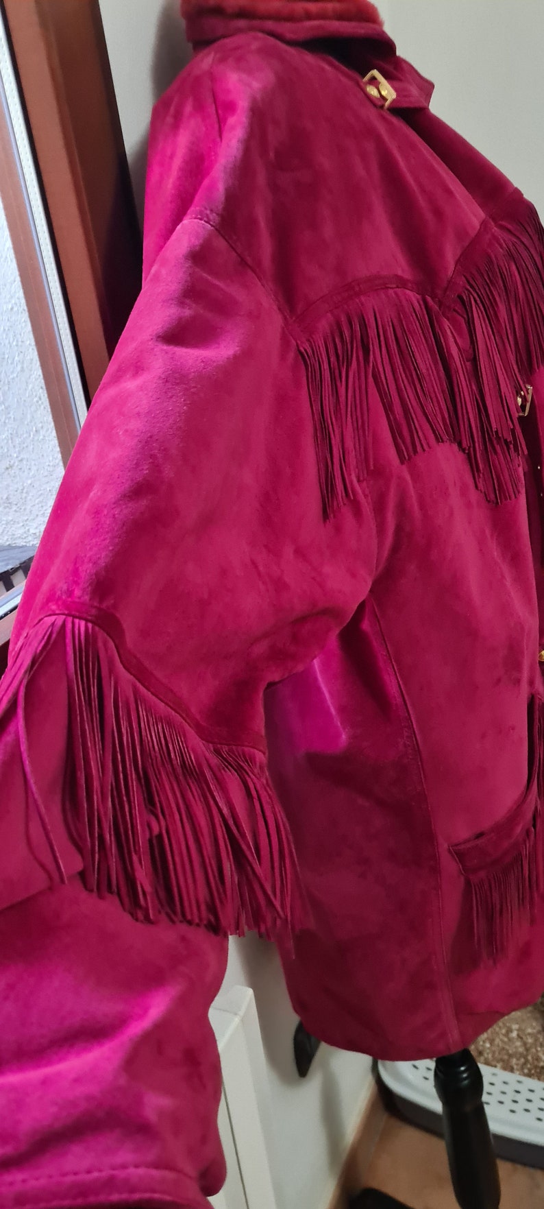 May include: A pink suede jacket with fringe detailing. The jacket has a gold buckle on the collar and a pocket on the front.