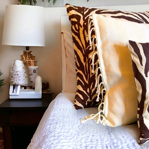 Op de afbeelding: Slaapkamer met kussens met een zebraprint in bruin en wit. Een crèmekleurig kussen met franjes is ook zichtbaar. Een witte lamp met een gouden voet staat op een donkerbruin nachtkastje.