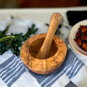 Könnte beinhalten: Ein Mörser und Stößel aus Olivenholz. Der Mörser ist eine tiefe Schüssel mit einem natürlichen Holzmaserungsmuster. Der Stößel ist ein glatter, abgerundeter Zylinder mit einem Griff.