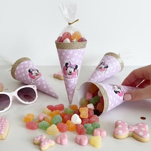 May include: Pink Minnie Mouse-themed party favors. The cone-shaped containers are filled with colorful candies and topped with gold glitter. Also includes heart and bow-shaped cookies and white sunglasses.