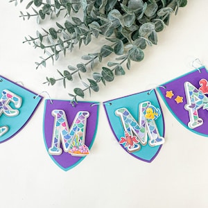 May include: A purple and blue banner with the name "EMMA" spelled out in silver letters with a mermaid scale pattern. Each letter has a mermaid or sea creature design on it.