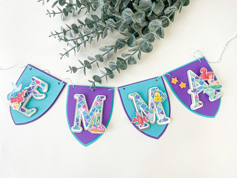 May include: A purple and blue banner with the name "EMMA" spelled out in silver letters with a mermaid scale pattern. Each letter has a mermaid or sea creature design on it.