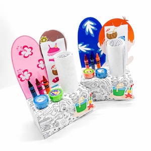 May include: Two surfboard-shaped craft kits with crayons and accessories. One is pink with white flowers, the other is blue with white leaf designs. Each kit includes a white cylinder, crayons, and small containers. The base is a black and white beach scene.