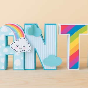 May include: Wooden letters spelling out the word "ANT". The letter "A" has a rainbow and cloud design. The letter "N" has blue and white stripes with cloud accents. The letter "T" has a rainbow design.