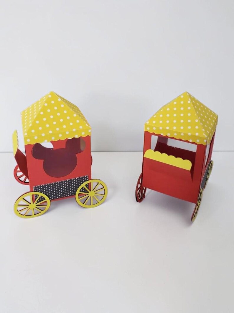 May include: Two red paper train carriages with yellow polka-dot roofs and yellow wheels. One carriage has a Mickey Mouse cutout. The carriages are on a white surface.