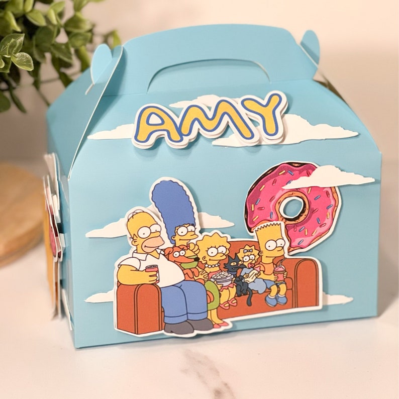 Puede incluir: Una caja de regalo azul y blanca con asa. La caja tiene una imagen de la familia Simpson en la parte delantera, con la palabra "AMY" en letras amarillas en la parte superior. La caja est&aacute; decorada con nubes blancas y una rosquilla rosa.