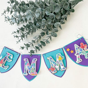 May include: A purple and blue banner with the letters "Emma" in white with a mermaid scale pattern. Each letter has a different mermaid character on it. The banner is strung with white string.