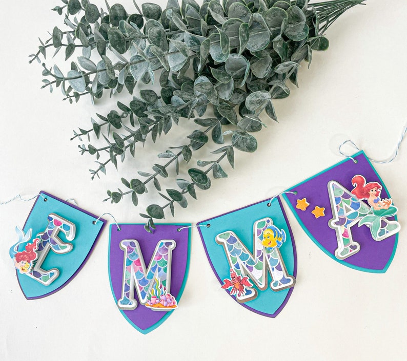 May include: A purple and blue banner with the letters "Emma" in white with a mermaid scale pattern. Each letter has a different mermaid character on it. The banner is strung with white string.