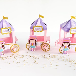 Op de afbeelding: Drie roze en paarse papieren uitsparingen met een circusthema. Elk heeft de vorm van een wagen met een tenttop, gouden wielen en een kleine vlag. Een stripmeisje met een roze hoed en blauwe jurk staat voor elke wagen. Gouden confetti is verspreid.