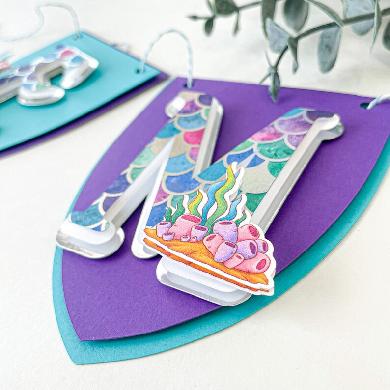 May include: A purple and teal banner with a white letter M decorated with a mermaid scale pattern and a coral reef design.
