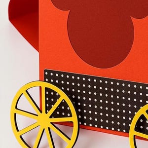 May include: A red and yellow paper craft resembling a Mickey Mouse-themed wagon. The wagon features a red body with a Mickey Mouse head cutout, yellow wheels, and a black and white polka dot accent.