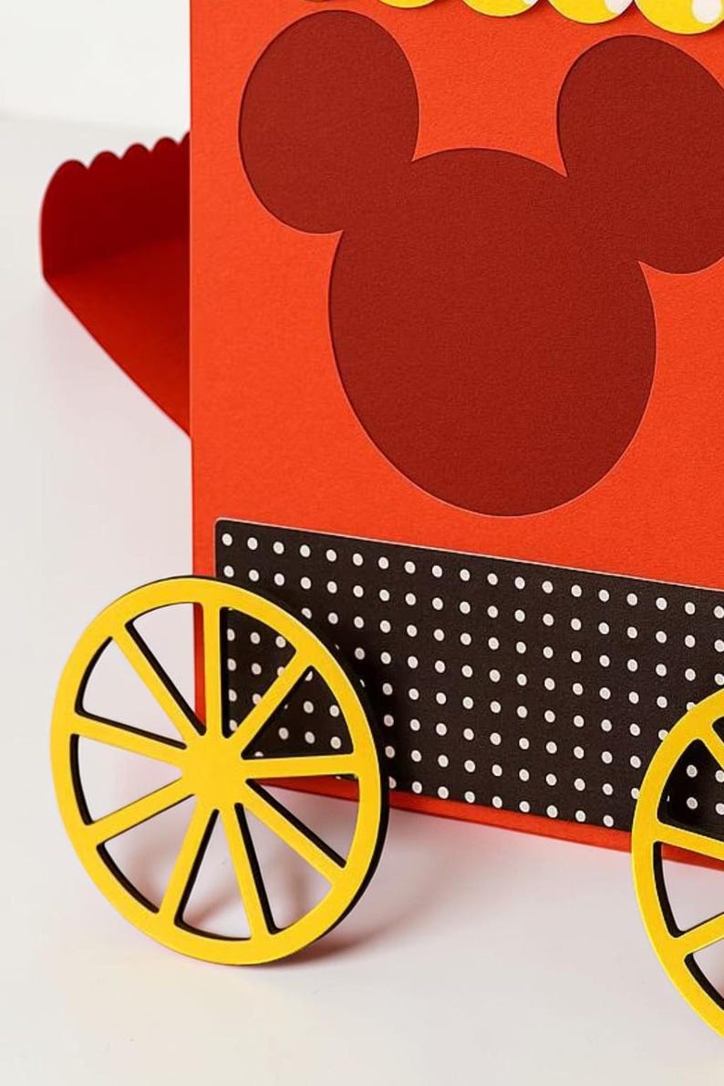 May include: A red and yellow paper craft resembling a Mickey Mouse-themed wagon. The wagon features a red body with a Mickey Mouse head cutout, yellow wheels, and a black and white polka dot accent.