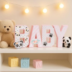 Pode incluir: Uma cena de berçário com a palavra "BABY" escrita em letras decorativas rosa e brancas. Um ursinho de pelúcia e um panda de pelúcia estão na prateleira. Abaixo estão três blocos de alfabeto.