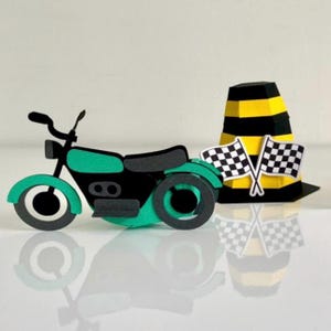 Op de afbeelding: Een papieren motorfiets in zwart en teal groen, met een zwarte zitting en wielen. Een zwart-gele kegel en twee geblokte vlaggen zijn ook aanwezig, wat wijst op een racethema. De objecten staan tegen een witte achtergrond.