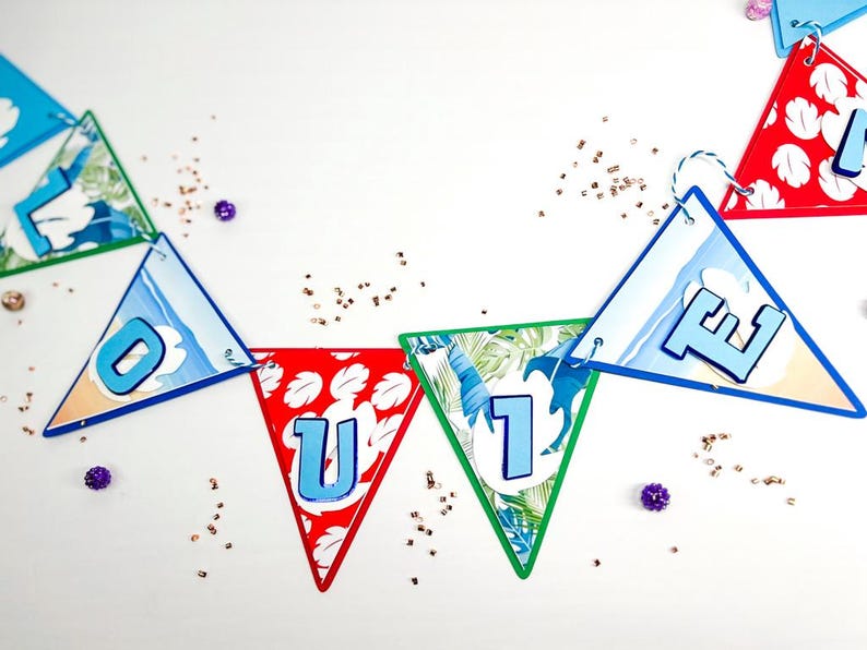 May include: A colorful paper banner with the word "OUTIE" spelled out in blue letters on triangular flags. Each flag has a different tropical design, including palm leaves, ocean waves, and flowers.