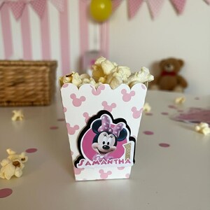 Op de afbeelding: Een witte popcornbox met een herhalend patroon van lichtroze muissilhouetten. De voorzijde toont een ronde uitsnede van een tekenfilmmuis met een roze strik, de naam SAMANTHA in roze letters en een gouden glitterende nummer één.