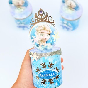 May include: A cylindrical container featuring a light blue snowflake pattern and a silver glitter trim. The top is adorned with a clear dome containing a character illustration, topped with a gold-colored crown cutout. The name Isabella is printed on the front.