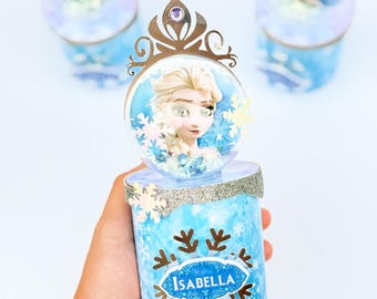 May include: A cylindrical container featuring a light blue snowflake pattern and a silver glitter trim. The top is adorned with a clear dome containing a character illustration, topped with a gold-colored crown cutout. The name Isabella is printed on the front.