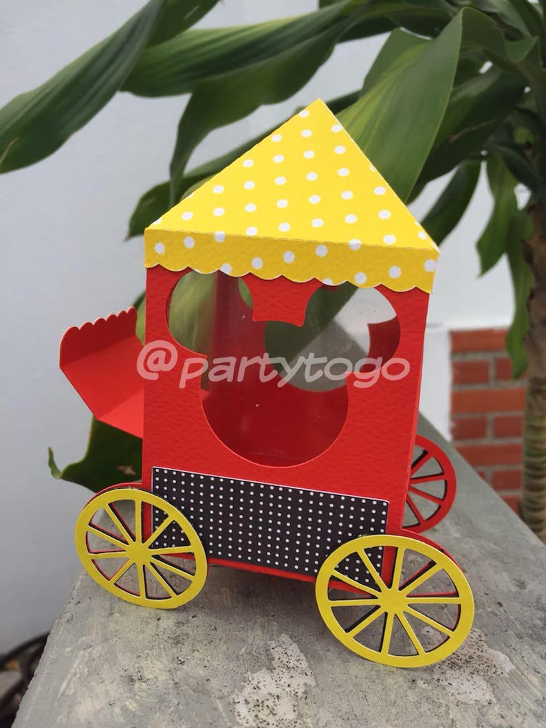 May include: A red and yellow paper train-shaped box with a Mickey Mouse head cutout. The train has yellow wheels, a yellow polka-dot roof, and a black and white dotted panel. The text "@partytogo" is visible on the side.