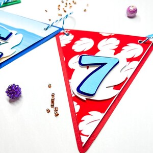 May include: A red triangular banner with a white floral pattern and a blue number 7. The banner is decorated with white and blue paper.
