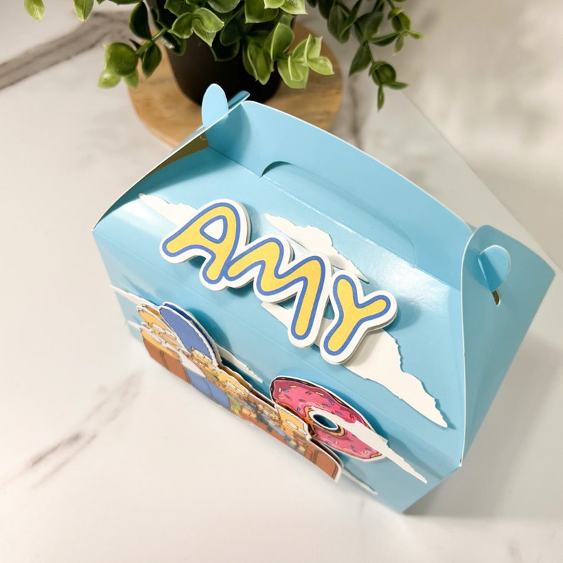 Puede incluir: Una caja de regalo azul con un fondo de nubes blancas y un nombre de dibujos animados amarillo "AMY" en la parte delantera. La caja presenta una imagen de dibujos animados de la familia Simpson con una rosquilla rosa.