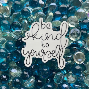 May include: White sticker with black lettering that reads "be kind to yourself" on a background of blue and clear glass stones.