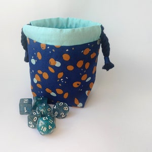 May include: A blue and orange fabric dice bag with a drawstring closure. The bag is filled with teal dice with white numbers.