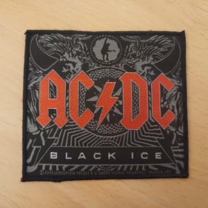 May include: A black and grey embroidered patch featuring the band name "AC/DC" in red lettering and the words "Black Ice" below. The patch includes a stylised lightning bolt and a design of two winged creatures.